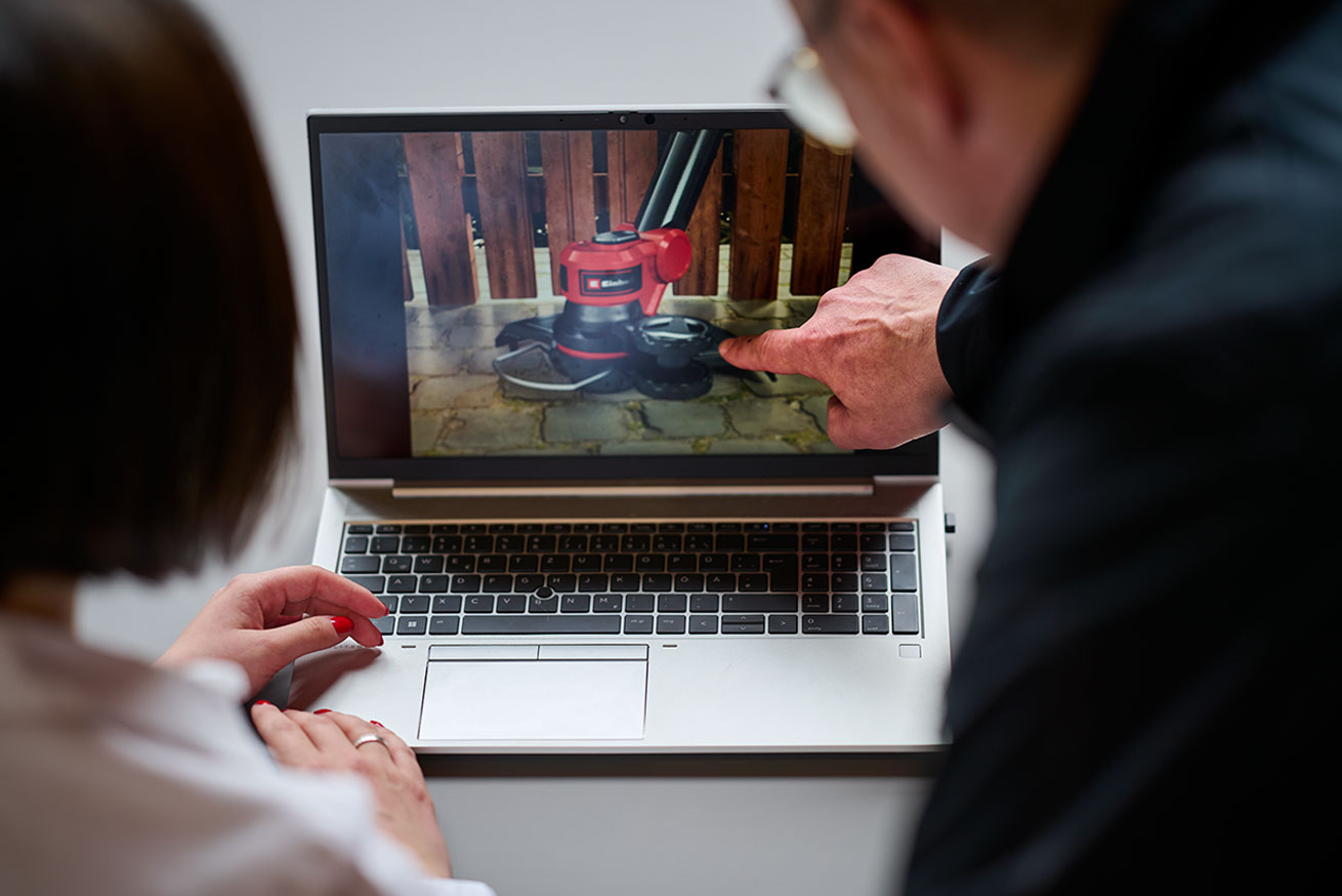 Two people are looking at a video or image of an Einhell floor-cleaning device on a laptop. One person is pointing at the screen.